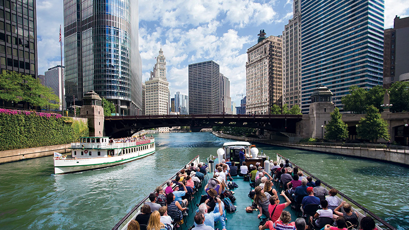 Two tour boats navigating a river in a bustling city, surrounded by modern skyscrapers and historic buildings, with greenery along the riverbanks and a steel bridge in the background
