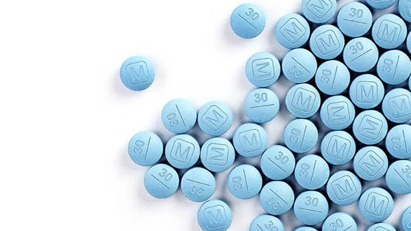 A close-up of numerous light blue, round pills scattered on a white surface. Each pill is marked with either an "M" inside a square or the number "30" with a line underneath.
