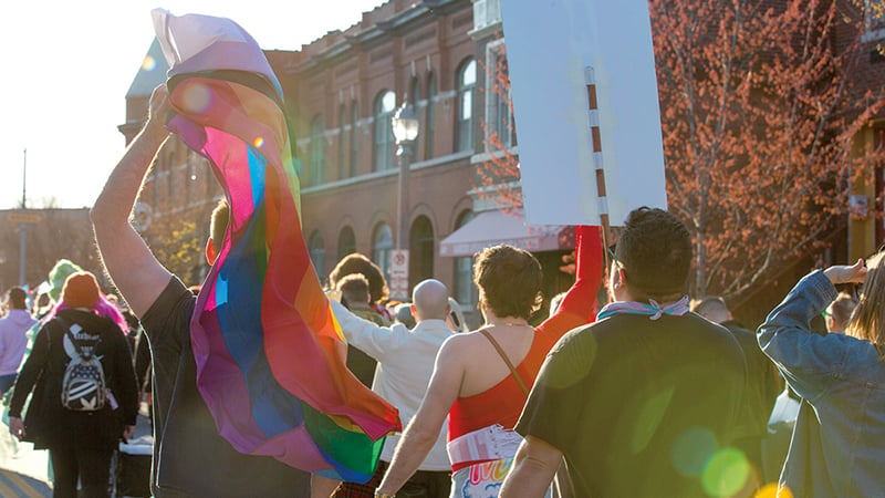 A vibrant Pride parade scene with a person holding a large rainbow flag aloft, surrounded by a crowd walking down a sunlit street.