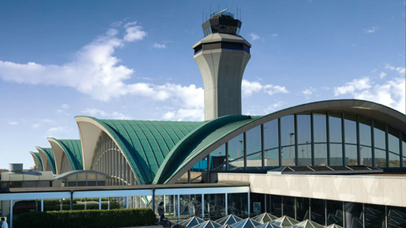 A modern airport terminal with curved green roofs and a tall air traffic control tower under a clear blue sky with scattered clouds.
