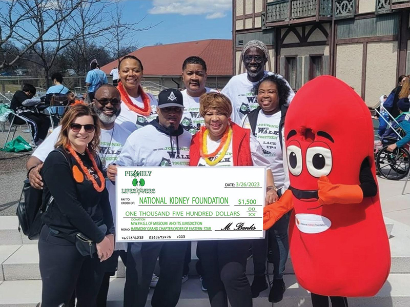 A diverse group of individuals, including a kidney-shaped mascot, holding a large novelty check for $1,500, presented to the National Kidney Foundation during an outdoor charity event.