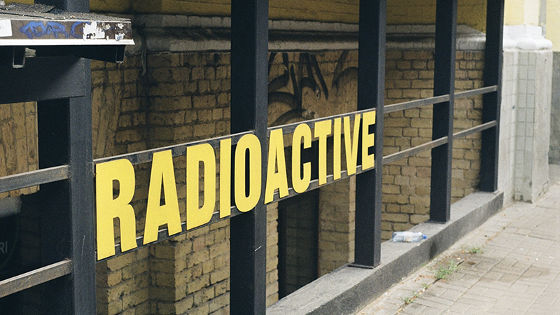 An urban entrance marked by a bold yellow "RADIOACTIVE" sign, surrounded by aged brick walls, graffiti, and a black metal railing.
