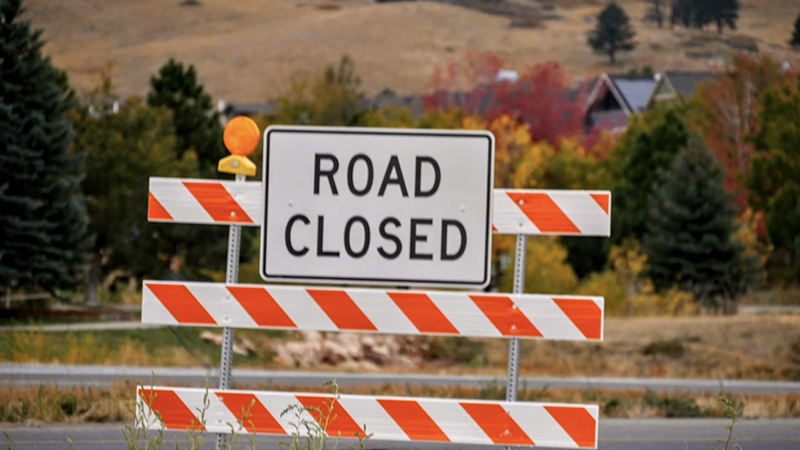 Striped road closed barrier with a flashing light blocking Highway 280, set against autumn trees and residential houses.