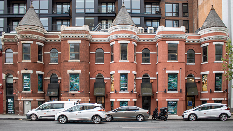 A red brick building with turret-like bay windows and arched upper-level windows, featuring "Retail Space Available" banners on the ground-floor storefronts. A modern dark-colored building with glass windows rises above it. Several vehicles, including a white van, compact cars, and a scooter, are parked on the street in front.