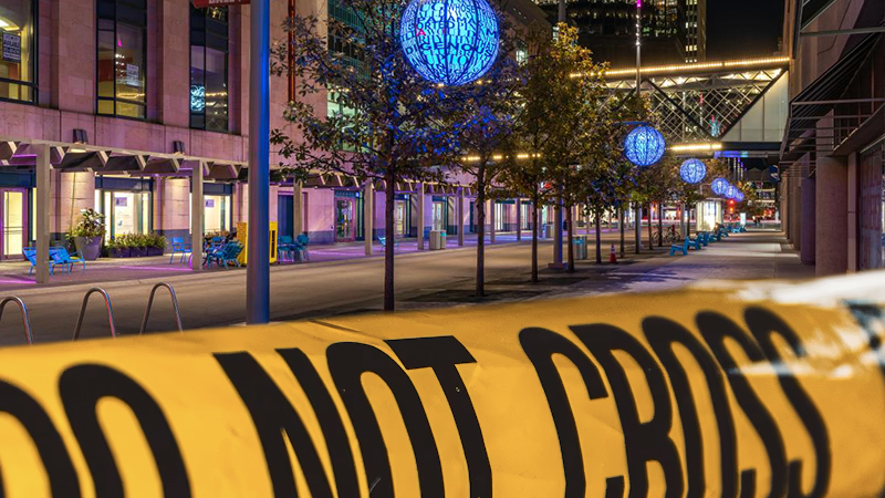 A nighttime view of Nicollet Mall in downtown Minneapolis, featuring glowing blue spherical lanterns hanging from trees, modern architecture, and a blurred yellow "DO NOT CROSS" tape in the foreground, symbolizing a restricted area.
