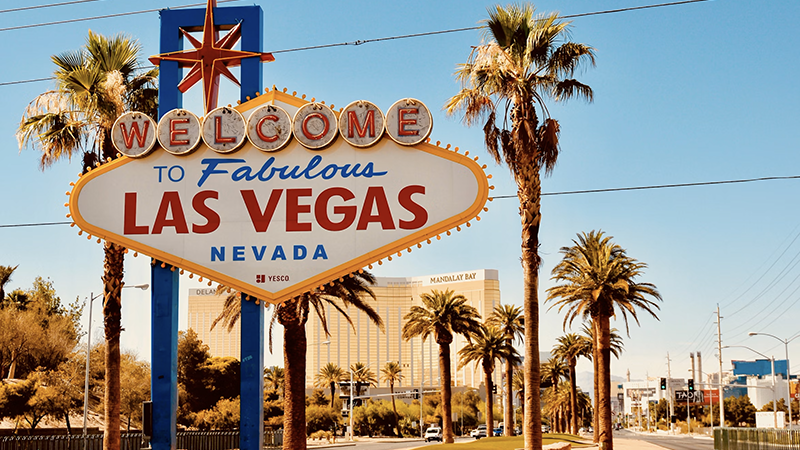 Welcome to Fabulous Las Vegas sign with palm trees and Mandalay Bay hotel in the background under a bright blue sky.