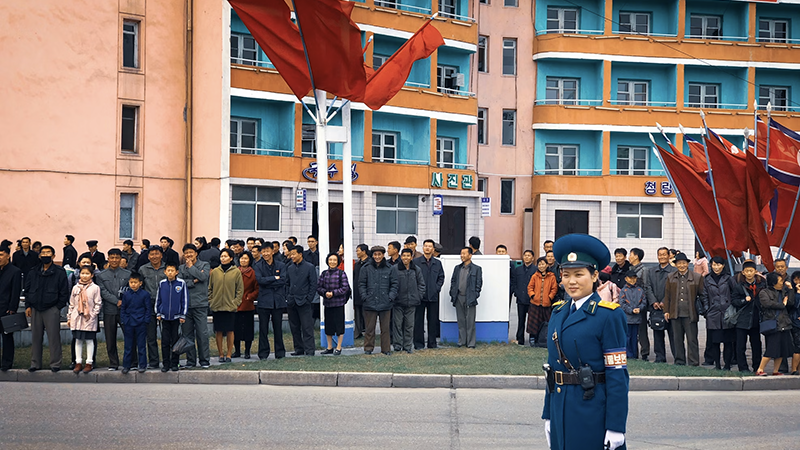A North Korean traffic officer stands in uniform, with a crowd and national flags in the background.
