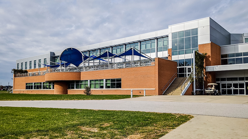 A modern multi-story building with a mix of brick and glass architecture, featuring a raised patio with blue fabric awnings, surrounded by a green lawn and a paved walkway under a cloudy sky.