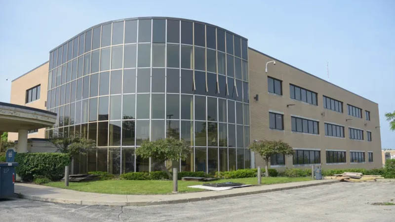 Three-story commercial building with a curved glass facade and beige brick exterior, surrounded by small trees and a paved parking area under a clear blue sky.
