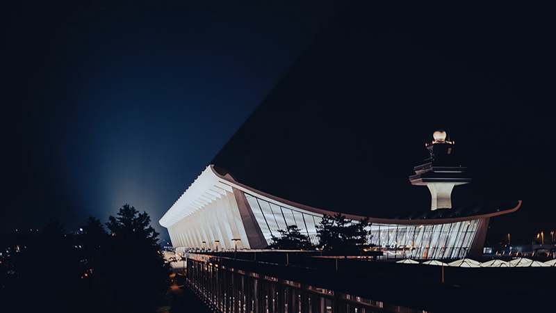 Nighttime view of Dulles International Airport's main terminal, showcasing its iconic curved roof and illuminated glass facade, with the control tower in the background."