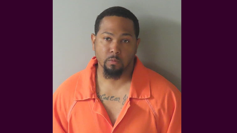 A mugshot of Cedric Hollimon wearing an orange jumpsuit, standing against a gray background with a neutral expression.