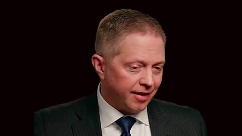 Headshot of Adam Fetcher, the former Chief Communications Officer for the City of Minneapolis, wearing a dark pinstripe suit and blue tie against a solid black background.