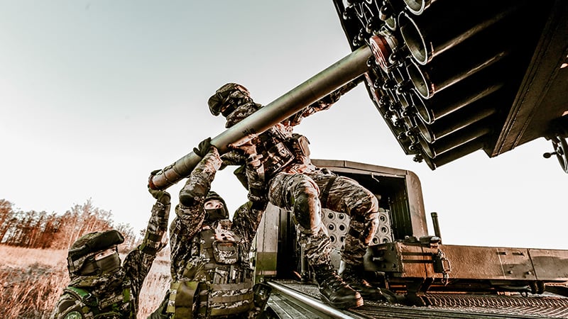 Soldiers in tactical camouflage uniforms loading a rocket into a multiple launch rocket system during field operations