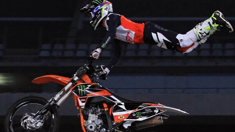 A dirt bike rider executing a high-flying aerial trick over a landing ramp inside a packed stadium.