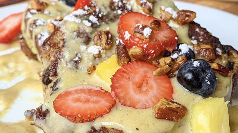 A close-up of a gourmet French toast dish topped with strawberries, blueberries, pineapple chunks, glazed pecans, powdered sugar, and a creamy yellow sauce, served on a white plate.