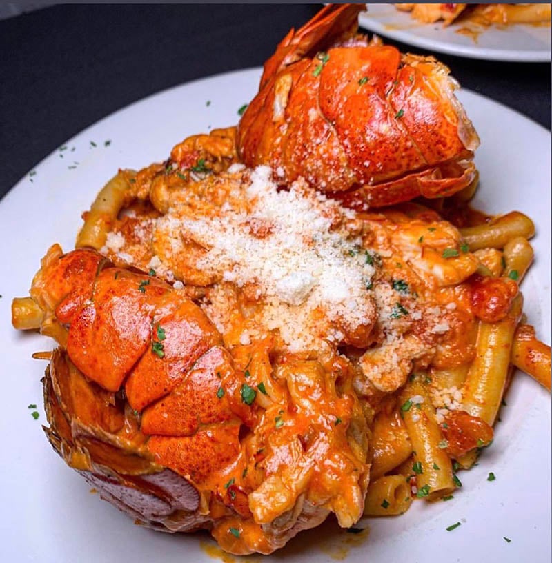 A white ceramic plate featuring a luxurious lobster pasta dish with two bright orange-red lobster tails, creamy tubular pasta in a rich red sauce, topped with grated cheese, breadcrumbs, and fresh herbs, set on a dark surface.