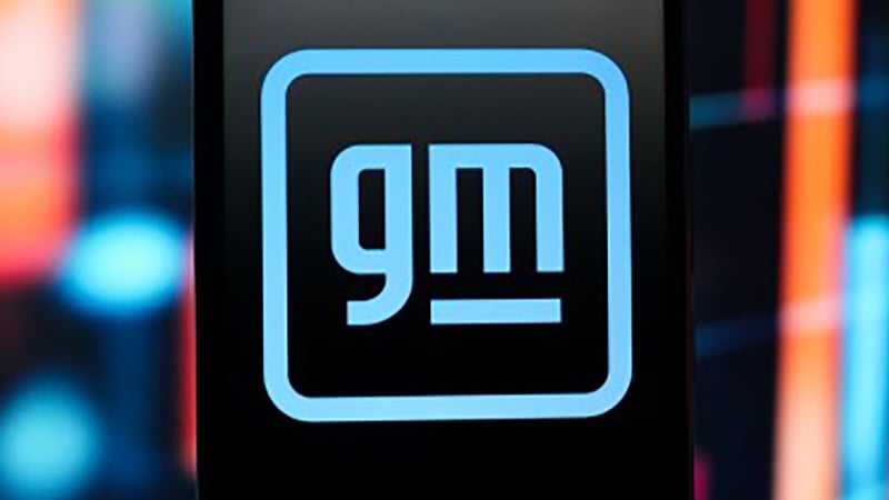 The General Motors logo in bright blue within a rounded square, set against a blurred, colorful background.