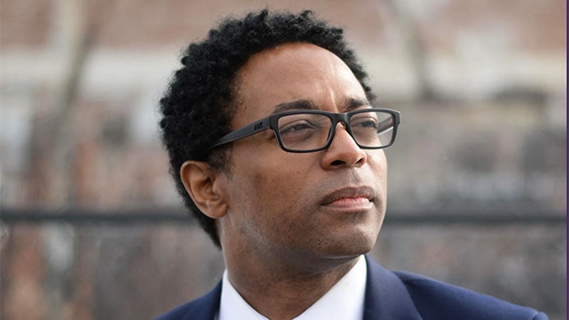 Wesley Bell in formal attire, looking contemplative, with a blurred urban background.