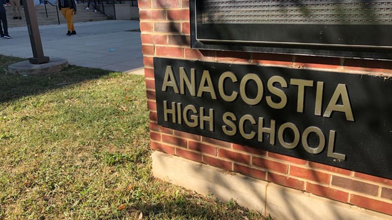 Anacostia High School sign on a red brick wall with a grassy foreground and people in the background.