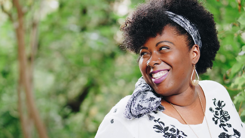 A smiling Black woman with a patterned headwrap and white embroidered top, standing outdoors with a blurred green background.
