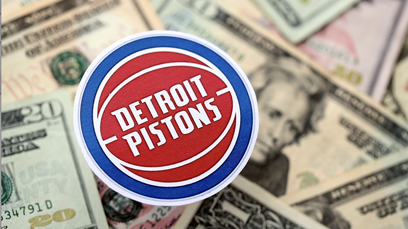 Detroit Pistons logo sticker placed on a pile of US twenty-dollar bills, symbolizing the financial aspects of sports and community.