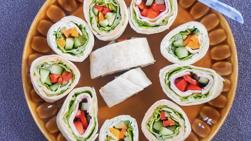 A plate of colorful tortilla pinwheels filled with fresh vegetables and deli meat, arranged on a decorative brown plate.