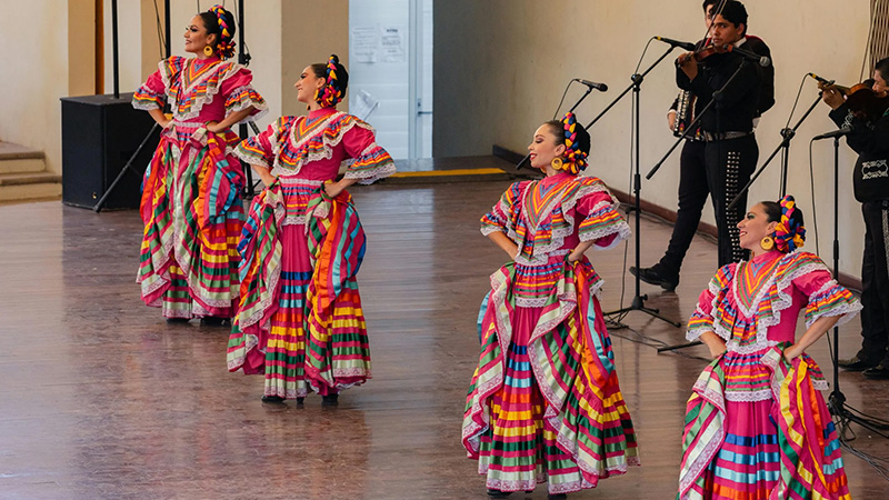 A group of dancers in traditional Mexican attire performing on stage, accompanied by a mariachi band, during a cultural celebration.