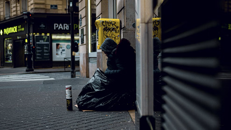A homeless individual huddled on a sidewalk in winter, wrapped in a tarp, with a pharmacy sign in the background.
