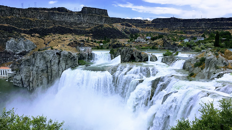 Panoramic view of a powerful waterfall cascading over rocky cliffs under a bright blue sky in Idaho.