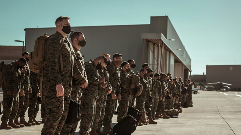 A group of military personnel in camouflage uniforms and face masks stand in a disciplined formation on an airfield, with a hangar and aircraft visible in the background under a clear blue sky.