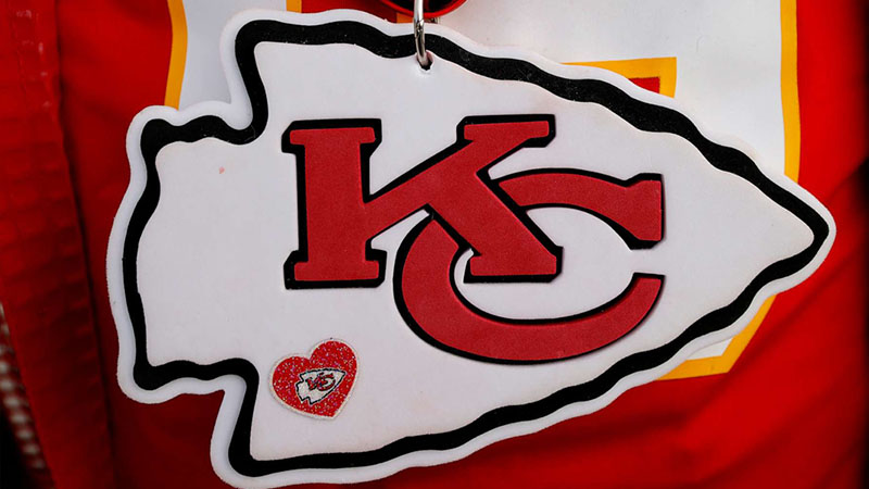 A white arrowhead-shaped ornament featuring the Kansas City Chiefs' red "KC" logo, with a glittery red heart embellishment, hanging against a blurred red and yellow background.