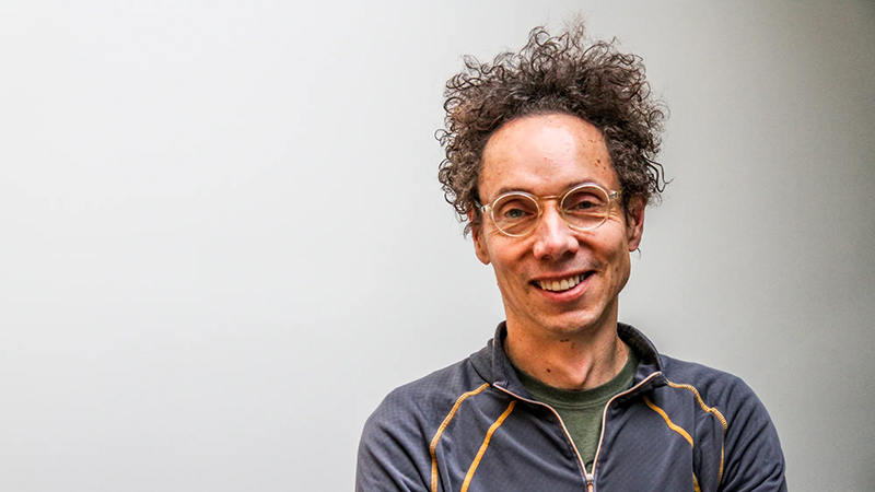 Malcolm Gladwell smiling in a medium close-up portrait, wearing glasses and a dark jacket, against a plain gray background.