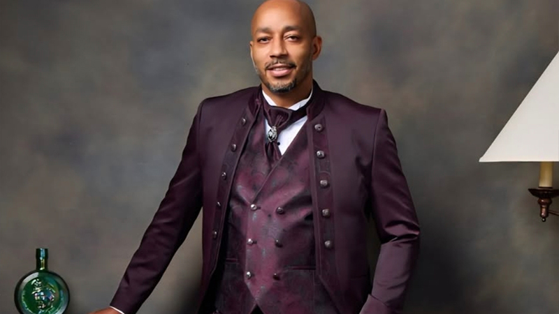Portrait of Newark artist Malik Whitaker in a maroon suit, smiling confidently, with a textured gray background.