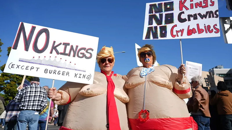 Protesters at the "No Kings" rally holding signs, dressed in costumes, and advocating for democracy under a clear blue sky.