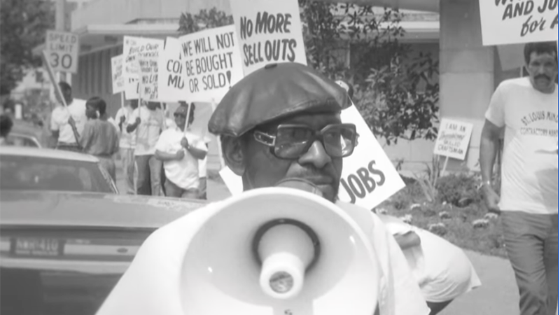 Percy Green speaking into a megaphone during a civil rights protest, surrounded by demonstrators holding signs advocating for equality.