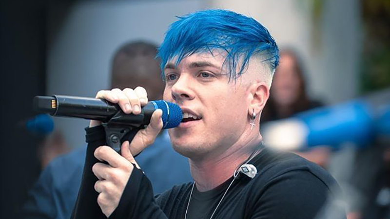 A performer with bright blue hair singing into a microphone during a live event, with a blurred audience in the background.