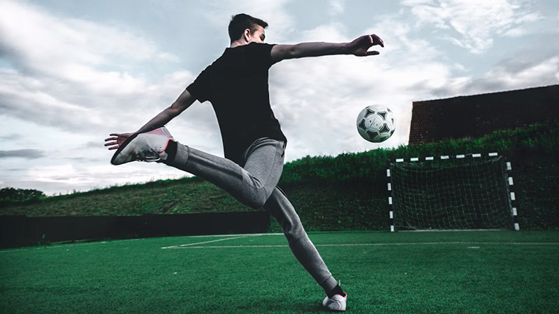 A young man kicking a soccer ball on a green field with a goal in the background, under a cloudy sky.