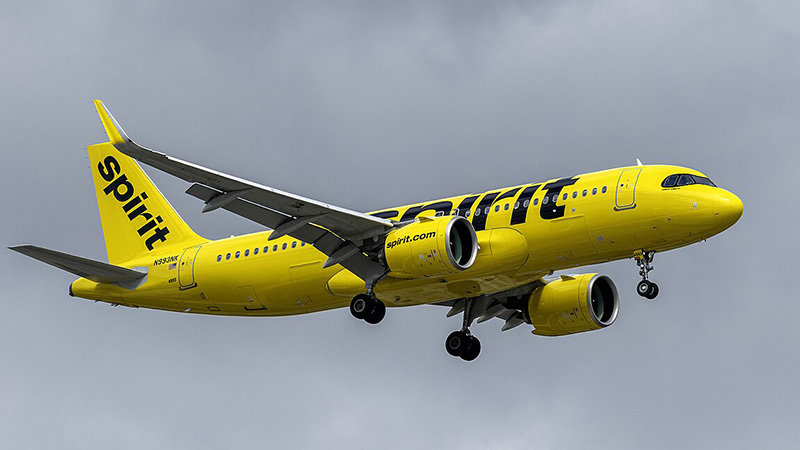 A bright yellow Spirit Airlines airplane in flight with landing gear extended, set against a cloudy sky.