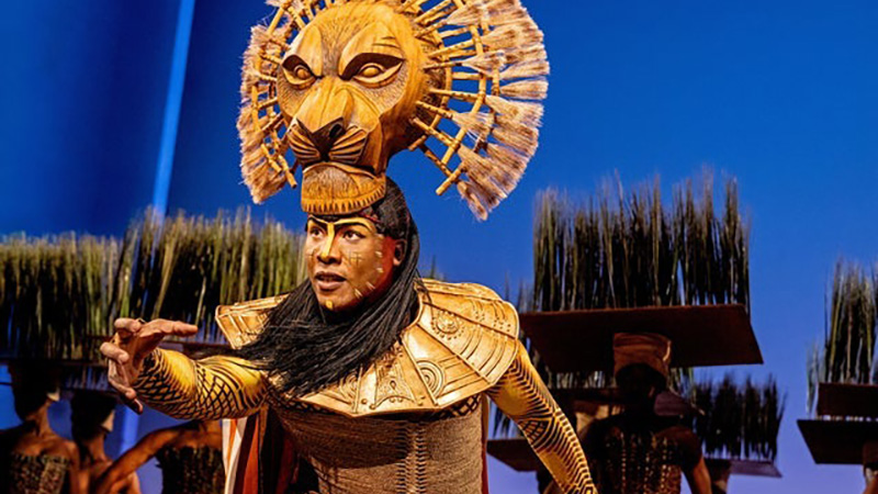A performer from The Lion King musical wearing an ornate lion headpiece and golden costume on stage with a blue backdrop and grass-like props.