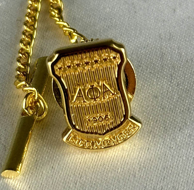 Polished gold Alpha Phi Alpha Life Member tie pin on dark suit lapel.