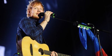 Ed Sheeran singing into a microphone with his acoustic guitar on stage during a stadium tour.