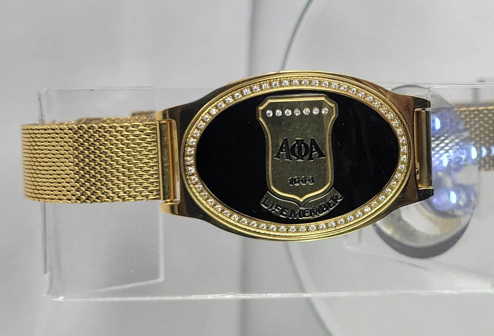 Studio display of the Alpha Black Life Member Mesh Band Bracelet with gold woven strap and crystal-encrusted black shield face