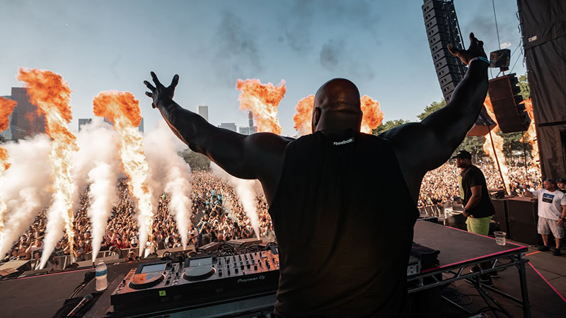 Rear view of a muscular DJ, seen from behind, standing on a large outdoor stage with arms raised and spread wide, facing a massive festival crowd. Multiple pyrotechnic flames and smoke bursts are erupting from the front of the stage.