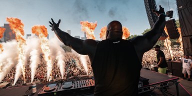 Rear view of a muscular DJ, seen from behind, standing on a large outdoor stage with arms raised and spread wide, facing a massive festival crowd. Multiple pyrotechnic flames and smoke bursts are erupting from the front of the stage.