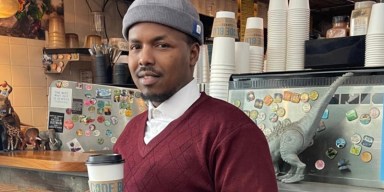 A portrait of a young Somali man wearing a grey beanie and maroon sweater vest, holding a paper coffee cup inside a local coffee shop.