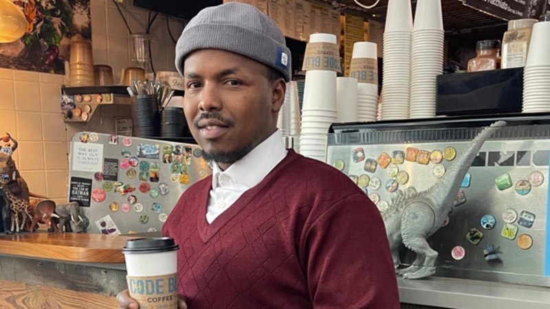 A portrait of a young Somali man wearing a grey beanie and maroon sweater vest, holding a paper coffee cup inside a local coffee shop.