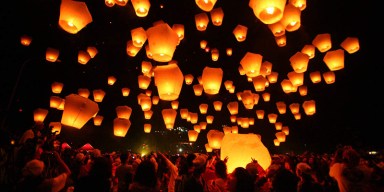 Glowing illuminated lanterns rising into the night sky over a crowd at an outdoor evening festival.