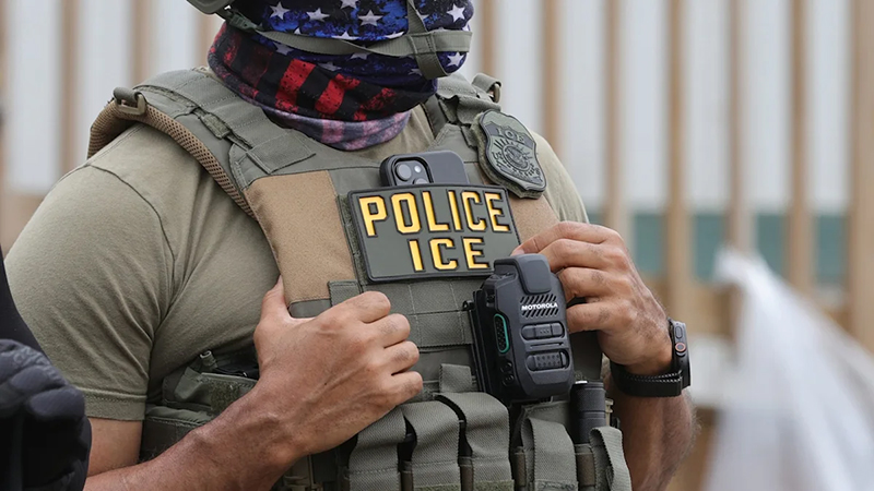Close-up of an ICE officer wearing a tactical vest with a Police ICE patch and body camera during an enforcement operation.