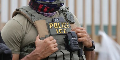 Close-up of an ICE officer wearing a tactical vest with a Police ICE patch and body camera during an enforcement operation.