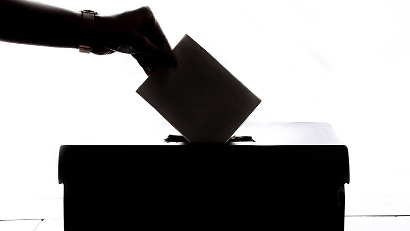 Silhouette of a hand placing a paper ballot into the slot of a ballot box against a bright white background.